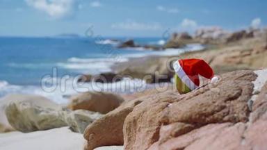 热带沙滩上的椰子上的圣诞帽，海浪在模糊的背景下滚动到岩石海岸线上